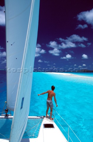 Man stands look out on the bow of a cruising catamaran approaching blue shallow water and a sandy sp
