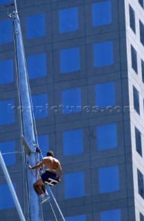 Crew member climbing mast of maxi yacht in front of office block