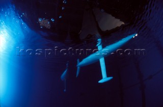 Model yachts racing in a blue swimming pool shot from underwater with lighting, showing keels and rudders