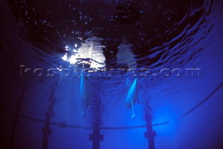 Model yachts racing in a blue swimming pool shot from underwater with lighting, showing keels and rudders