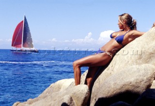 Girl leaning against rocks looking out to sea at yacht