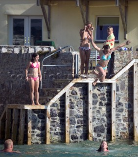 Children and kids jump and dive into the water from a wall in front of a luxury house in Salcombe Harbour