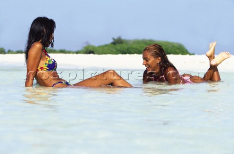 Two girls in bikinis lying in shallow water on sandy beach 