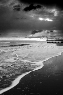 Black and white image of sea lapping onto a hard sand beach with groyne in distance