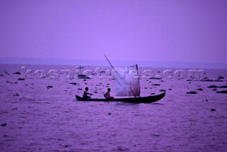 Two men sailing a canoe with a homemade sail at dusk in the Kerala Backwaters, India.