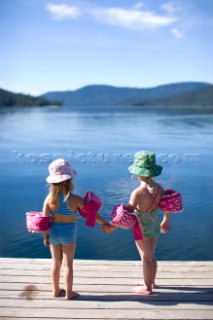 Della Rose Wheatcroft and her friend Sofia Platte stand at the end of a dock holding hands with their bathing suits, sun hats and water wings on. They are on vacation at Priest Lake, Idaho.