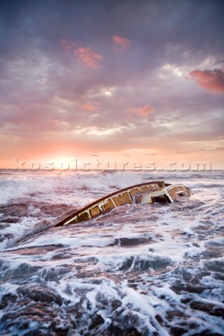 Waves crash over the remains of a sailboat with a dramatic sunset in the background  The sailboat th