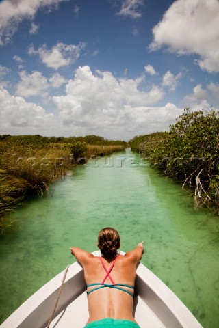 A young woman in a bikini sits in the bow of a white boat as it moves through tealgreen water of the